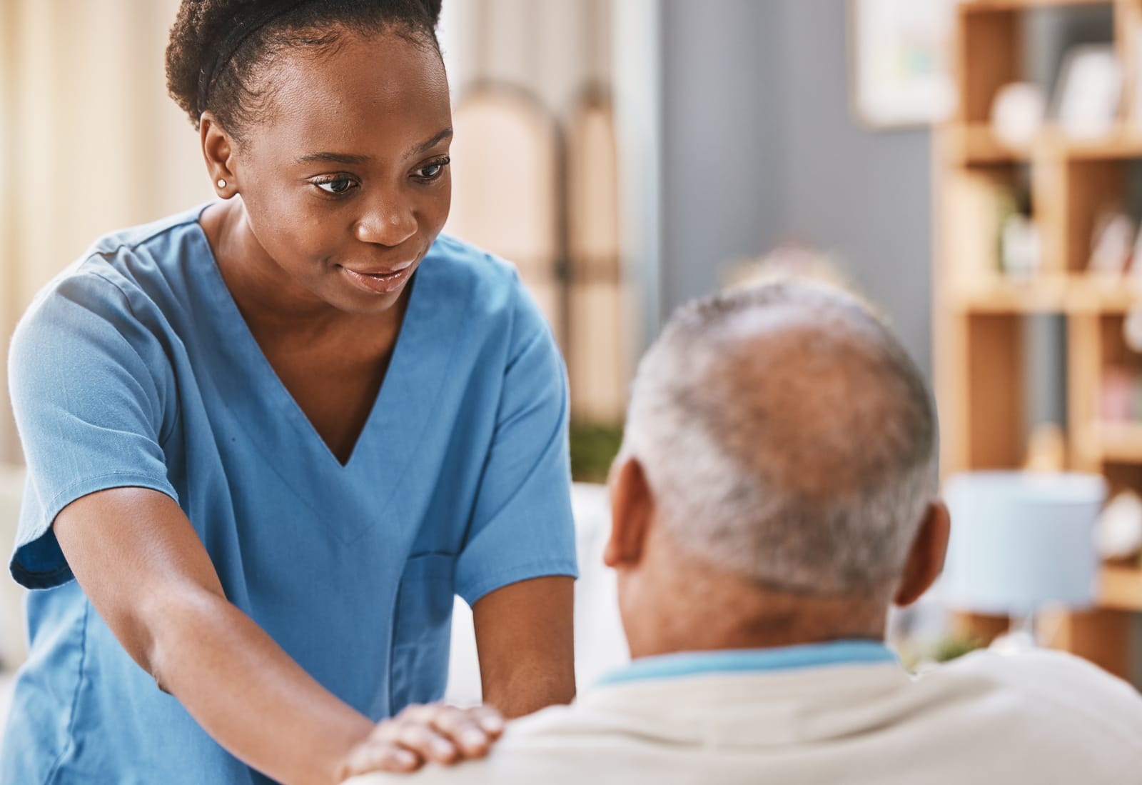 A care professional in scrubs attentively supporting an older client in a warm home setting