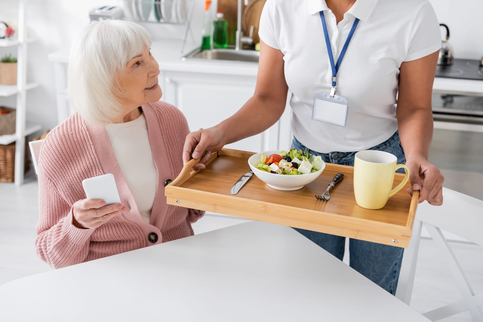 A care worker with an ID badge bringing a healthy meal on a tray to a smiling senior woman in her kitchen