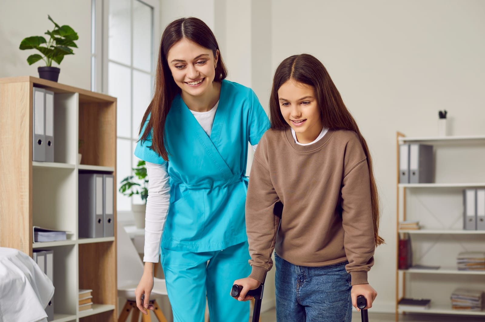 A caregiver supporting a smiling young girl as she walks with crutches